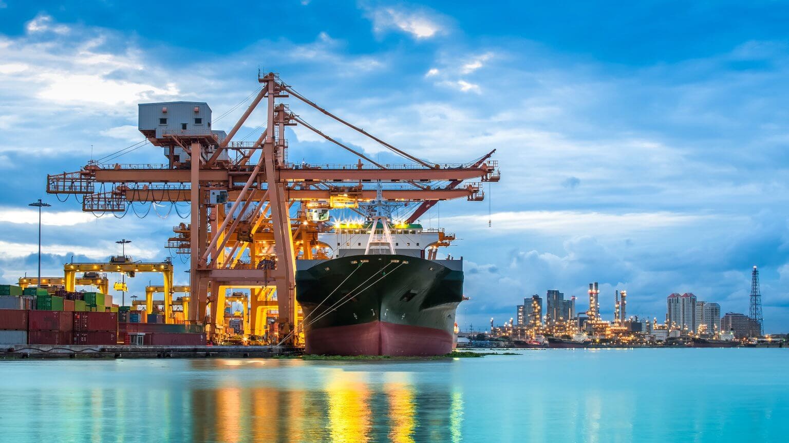 A modern container ship docked at an industrial port, with large cranes, shipping containers, and a cityscape with industrial buildings in the background under a cloudy blue sky.